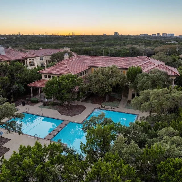 Aerial view of a luxury apartment complex featuring two swimming pools surrounded by greenery at sunset.