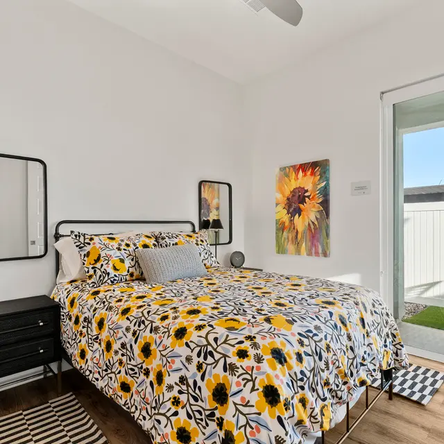 A bright bedroom featuring a bed covered in a floral yellow and white comforter. There's a small nightstand with a lamp, a framed mirror on the wall, and a large window with a view of a balcony. A colorful painting of flowers hangs on the wall.