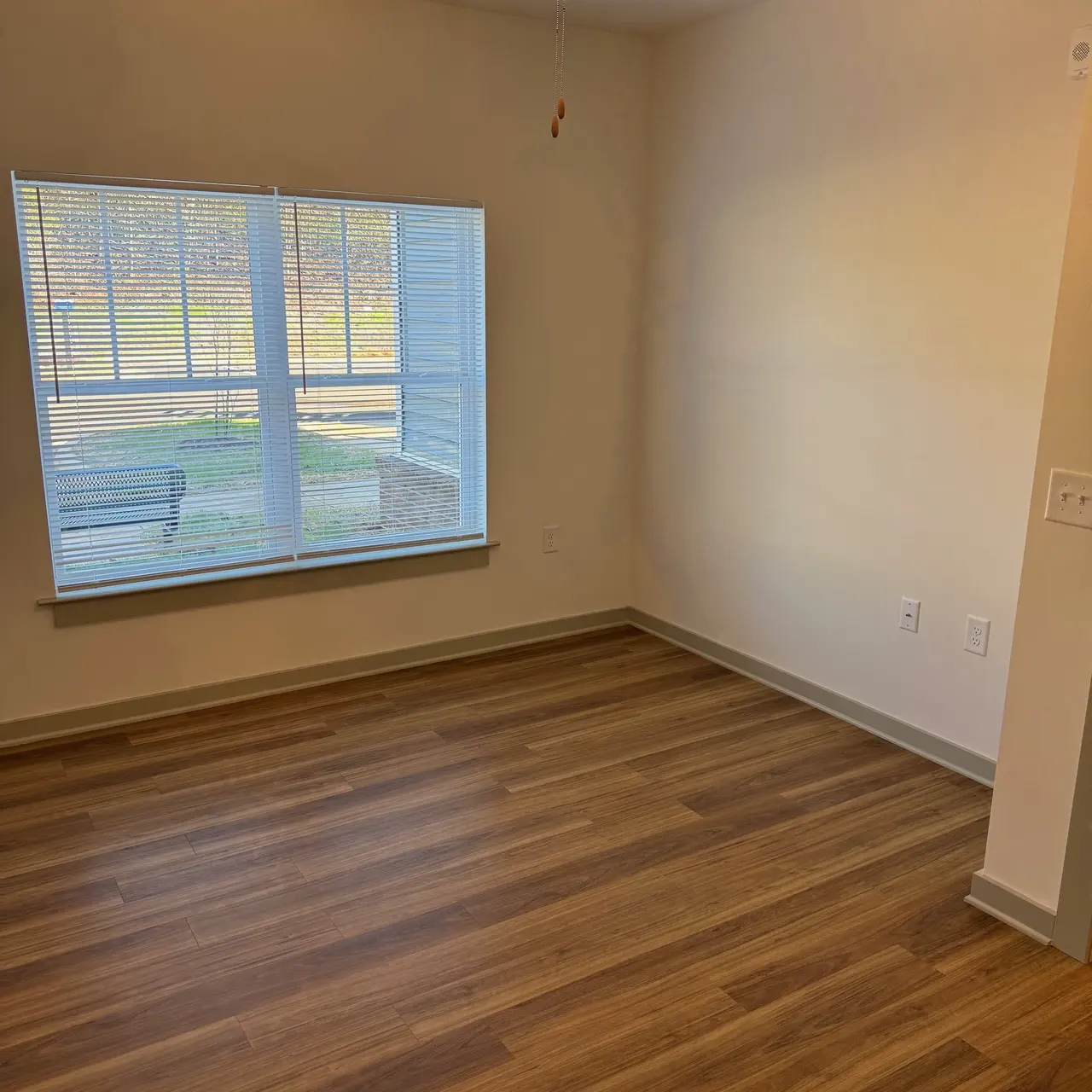 An empty room with a ceiling fan and a large window showcasing blinds. The floor is wooden with light brown tones, and sunlight is coming in through the window, illuminating one corner of the room.