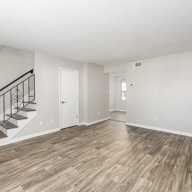 A spacious, empty living room with light-colored walls and wood-style flooring. A staircase with a black railing is visible on the left. A doorway leads to another room in the background.