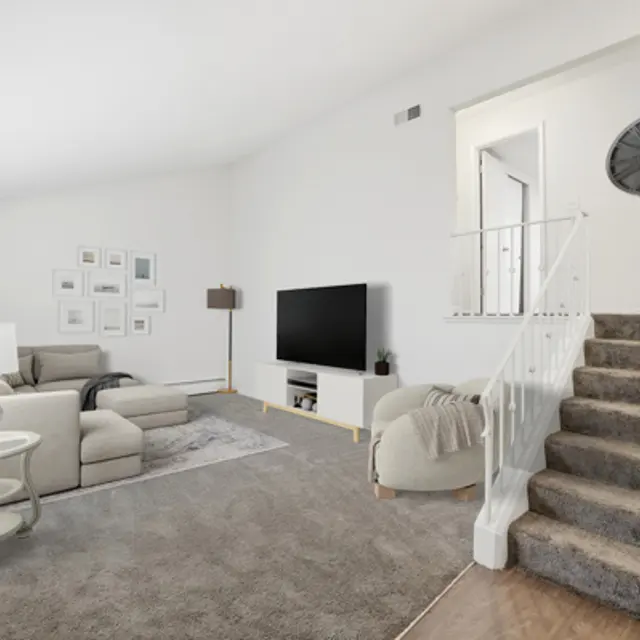 A bright, modern living room featuring a beige sectional sofa, a lamp, and a large TV opposite a decorative wall. There are stairs leading to another level, and light-colored walls with a few decorative elements.