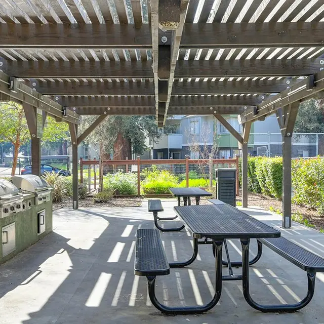 An outdoor pavilion featuring a pergola structure with several picnic tables and a grill station, surrounded by greenery and a tennis court.