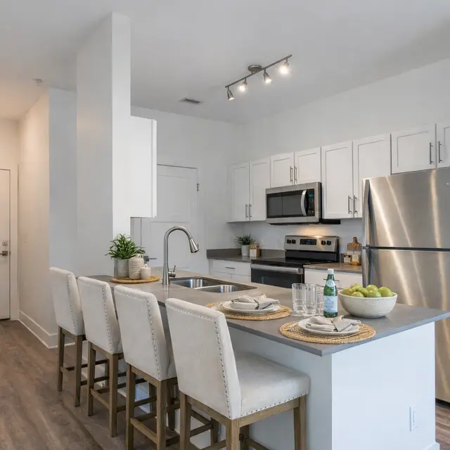 A modern kitchen featuring a large island with four bar stools, stainless steel appliances, and white cabinetry. The design is contemporary with clean lines and natural light.