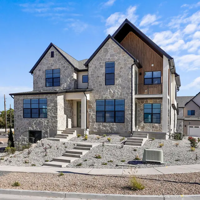 A modern two-story house with a stone facade and wooden accents, featuring large windows, landscaped grounds, and a clear blue sky in the background.