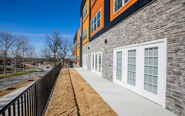 A modern apartment exterior with a stone facade and orange accents, featuring multiple glass doors leading to a patio area.