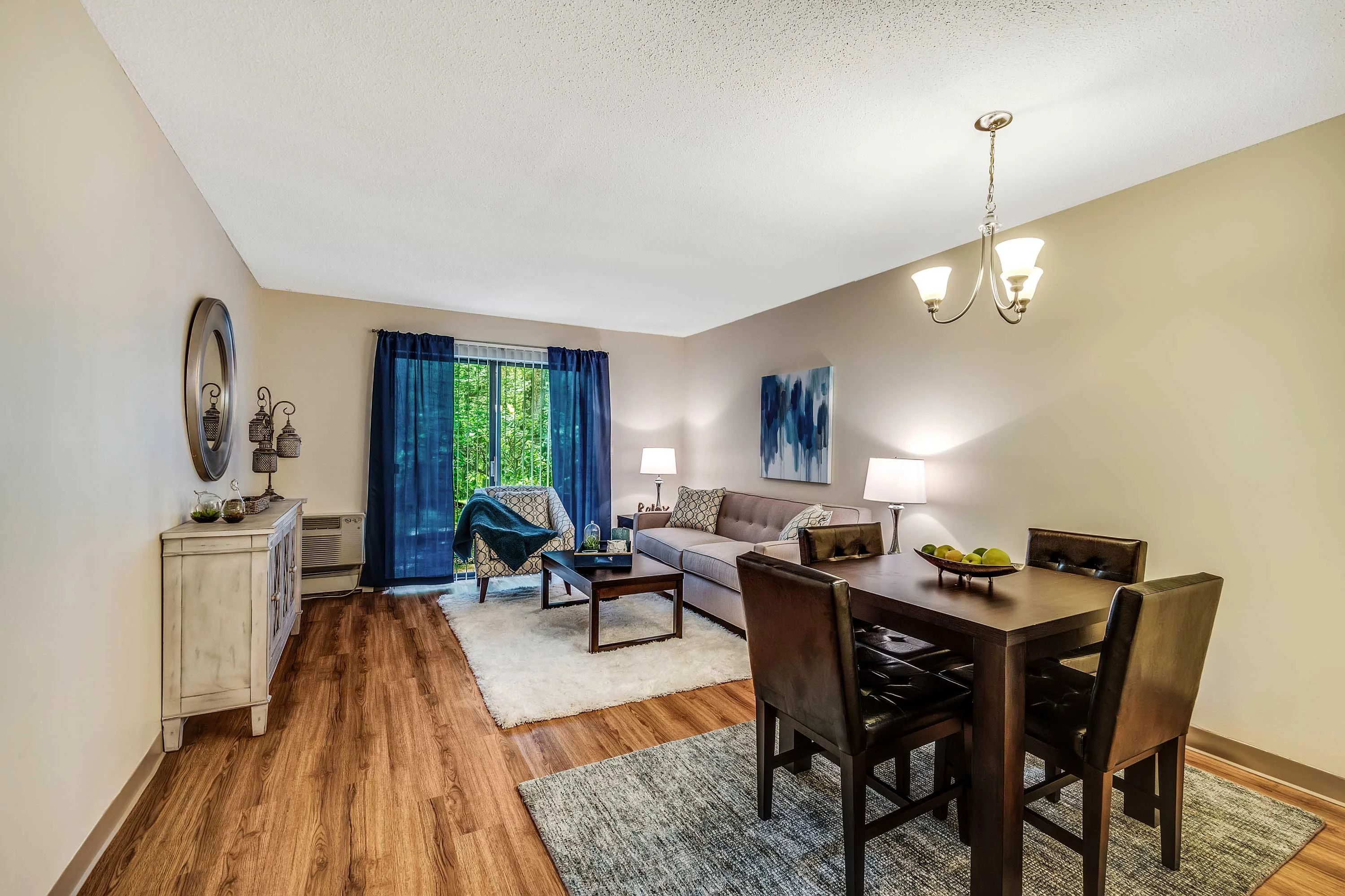 A modern living room with a dining area, featuring a sofa, dining table with chairs, and large windows with blue curtains.