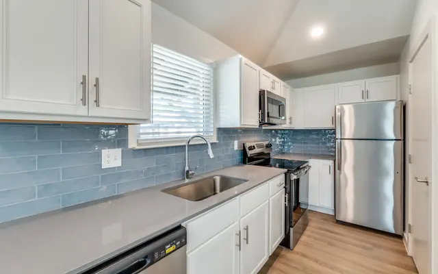Modern Kitchen Design A modern kitchen featuring white cabinetry, stainless steel appliances, and a blue backsplash. The kitchen includes a sink, stove, and refrigerator, with wooden flooring and natural light coming from a window.
