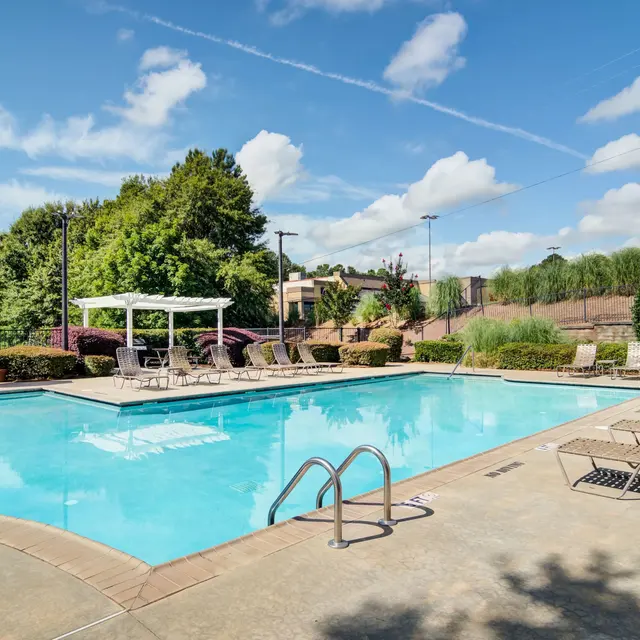 A bright outdoor swimming pool area with lounge chairs and green landscaping.