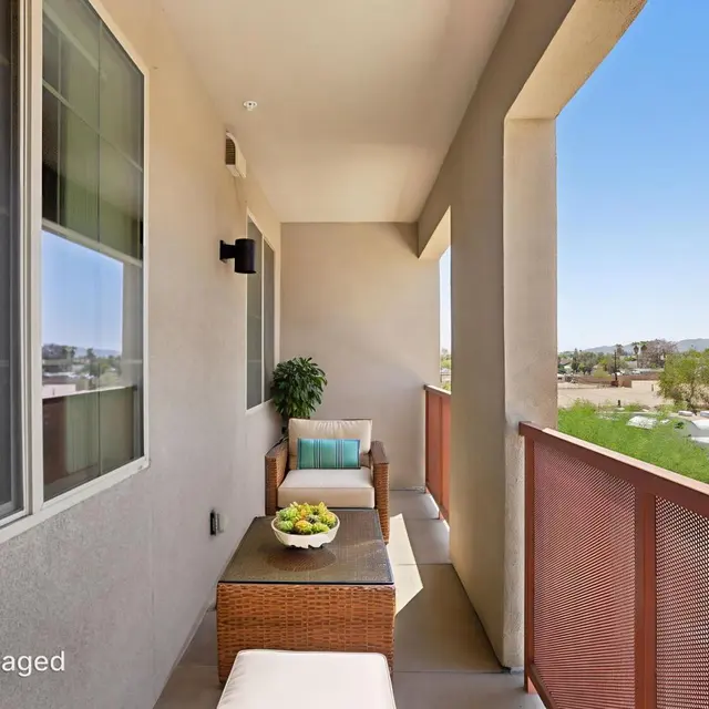 Modern Balcony with Scenic View A modern balcony featuring a small seating area with a wicker couch, coffee table, and decorative fruit bowl. The view includes distant mountains and greenery under a clear blue sky.