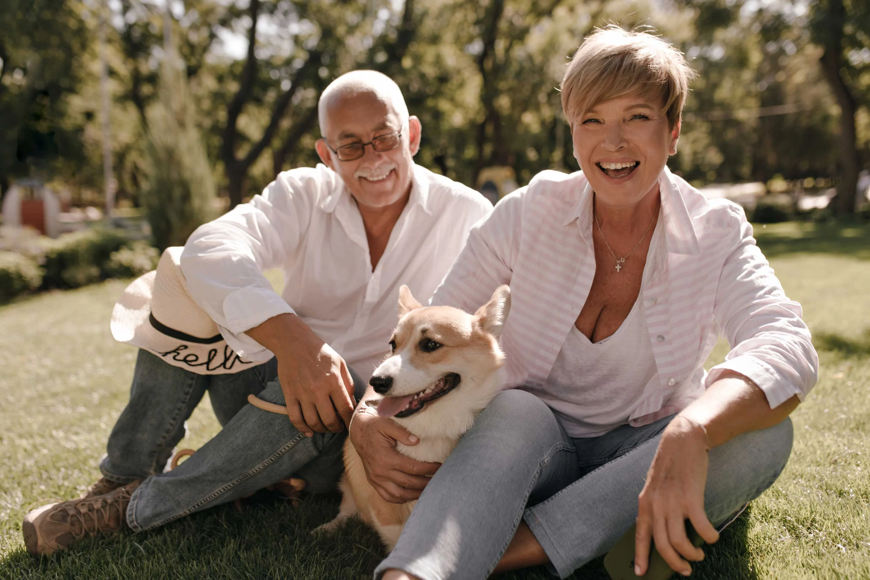 Couple Enjoying Time with Their Corgi A couple sitting on a grassy field, smiling and enjoying their time with a corgi dog. The woman is wearing a light pink shirt and the man is in a white shirt, both looking relaxed and happy.