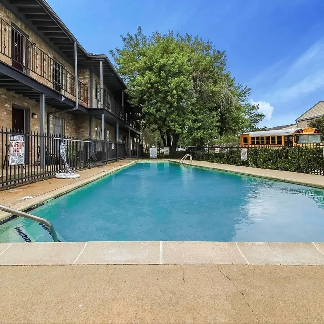 A clear swimming pool surrounded by stone pavement and a fence, next to a small apartment building featuring balconies. Lush trees and a school bus can be seen in the background under a bright blue sky.