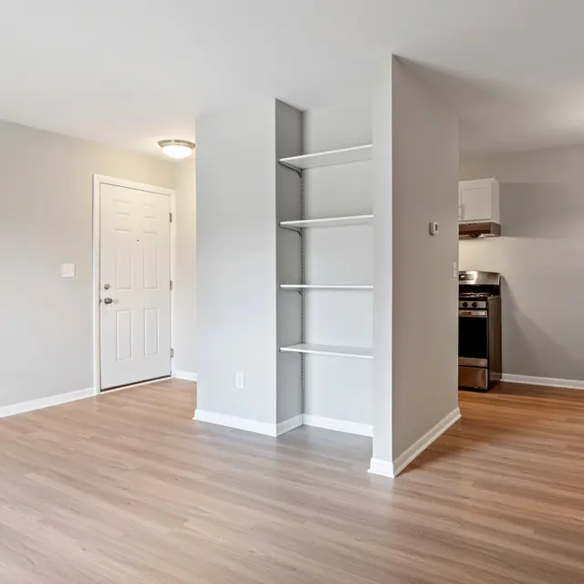 A spacious, modern apartment interior featuring wooden flooring, painted walls, shelves on a dividing wall, and kitchen appliances visible in the background.