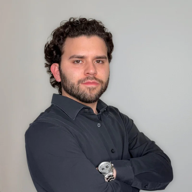 Portrait of a Man in Black Shirt A man with curly dark hair and a beard stands confidently with his arms crossed, wearing a black shirt and a watch on his left wrist, in front of a light gray background.