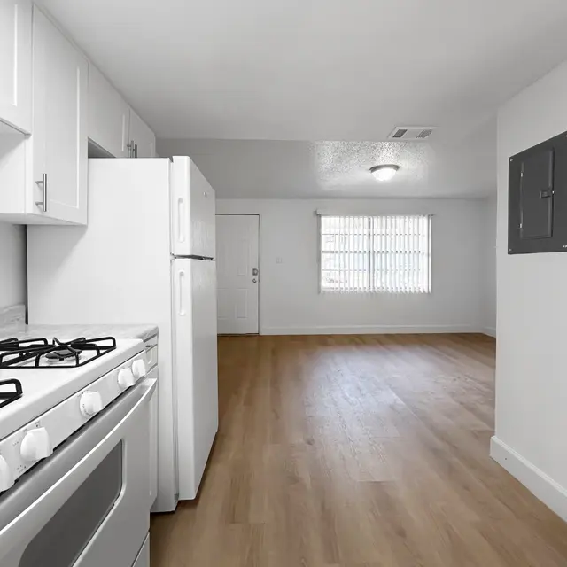 Small Apartment Interior An interior view of a small apartment showing a kitchen area with a gas stove and a refrigerator. The room has light wooden flooring, white walls, and a window with blinds.