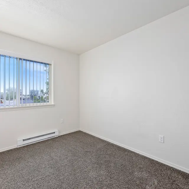 Bright Empty Room An empty room featuring beige carpet flooring and white walls. A window with vertical blinds lets in natural light, revealing an exterior view with trees and buildings in the distance.