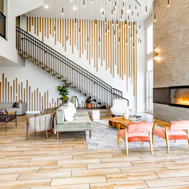 A modern living room featuring a stylish staircase, a decorative wooden wall, and an inviting fireplace. The space includes various seating options including a green sofa, pink chairs, and a wooden coffee table, with decorative plants adding to the aesthetic.