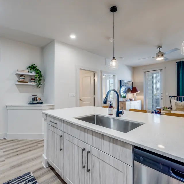A modern kitchen and living room area featuring a light-colored kitchen island, stainless steel appliances, and a comfortable seating area with natural light coming through large windows.