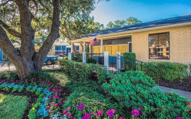 A charming house exterior with vibrant flowers in the foreground and lush greenery, featuring an American flag on the porch, and a clear blue sky.
