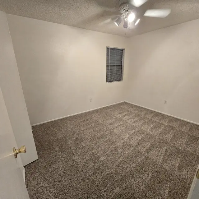 Empty Room Interior An empty room with beige carpet, light-colored walls, and a ceiling fan. There is a window with vertical blinds allowing some light in. A door is partially open, revealing the entrance to the room.