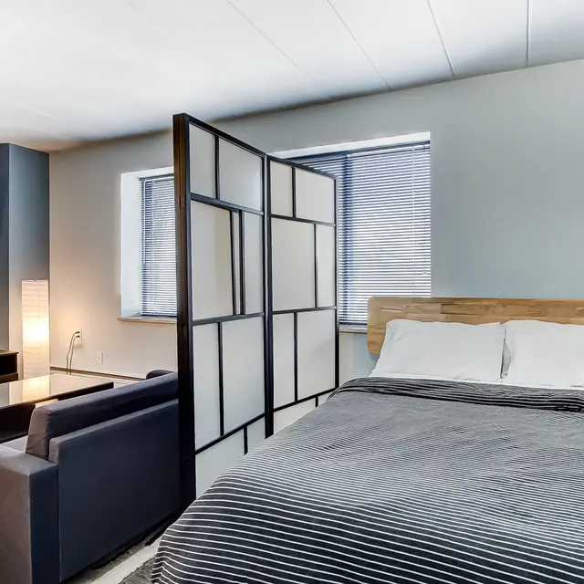 A modern bedroom featuring a bed with a wooden headboard, separated by a decorative screen. A cozy sitting area with a black couch is visible, along with a TV on the wall and a lamp illuminating the space.