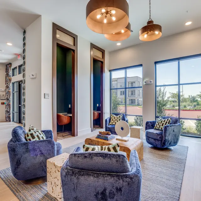 A contemporary living room with plush blue chairs, a stylish coffee table, and large windows allowing natural light. The decor includes pendant lights and a modern rug.