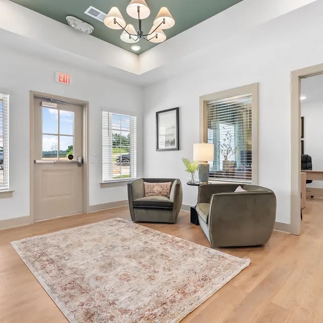 A modern lobby area with two armchairs, a small table with a lamp, and a large rug on wooden flooring. There are large windows letting in natural light and a doorway leading to another room.