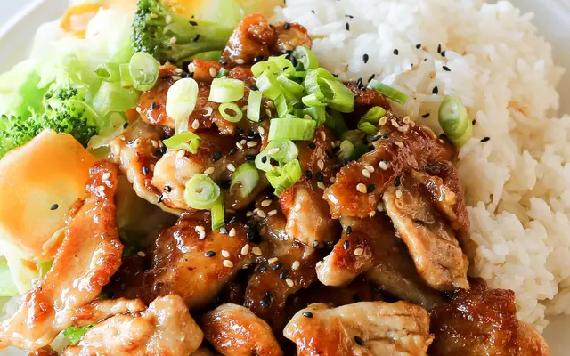 A plate of chicken pieces glazed in a dark sauce topped with chopped green onions and sesame seeds, accompanied by white rice and steamed vegetables, including broccoli and a leaf of lettuce.