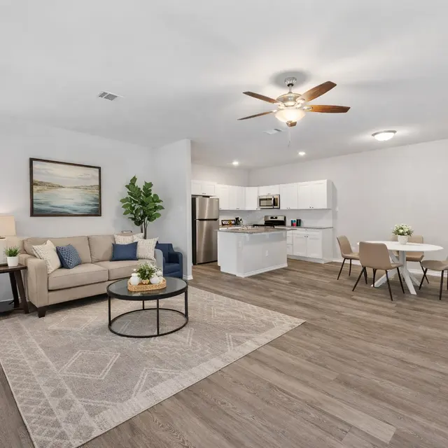 A modern open-concept living space featuring a cozy beige couch, a dining area with four chairs, and a kitchen equipped with stainless steel appliances. A welcome sign and decorative plants add to the homey atmosphere.