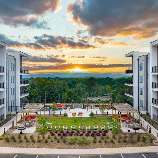 Aerial view of modern apartment buildings with a landscaped courtyard at sunset. The setting sun casts warm hues over the horizon and greenery.