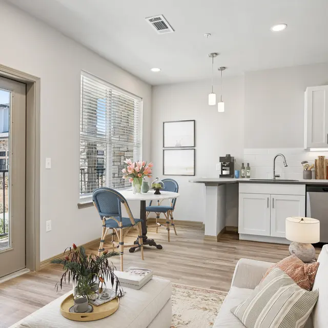 Cozy Modern Apartment Interior Interior of a modern apartment with a cozy living area and kitchenette. Features a small dining table with flowers, a sofa, and large windows letting in natural light.