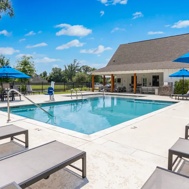 Outdoor Swimming Pool with Lounge Area A sunny outdoor swimming pool area featuring blue umbrellas, lounge chairs, and a house in the background.