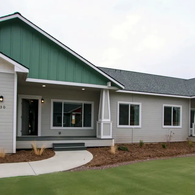 A modern single-story house featuring a combination of gray and green siding with a large front porch, large windows, and a driveway leading to a garage.