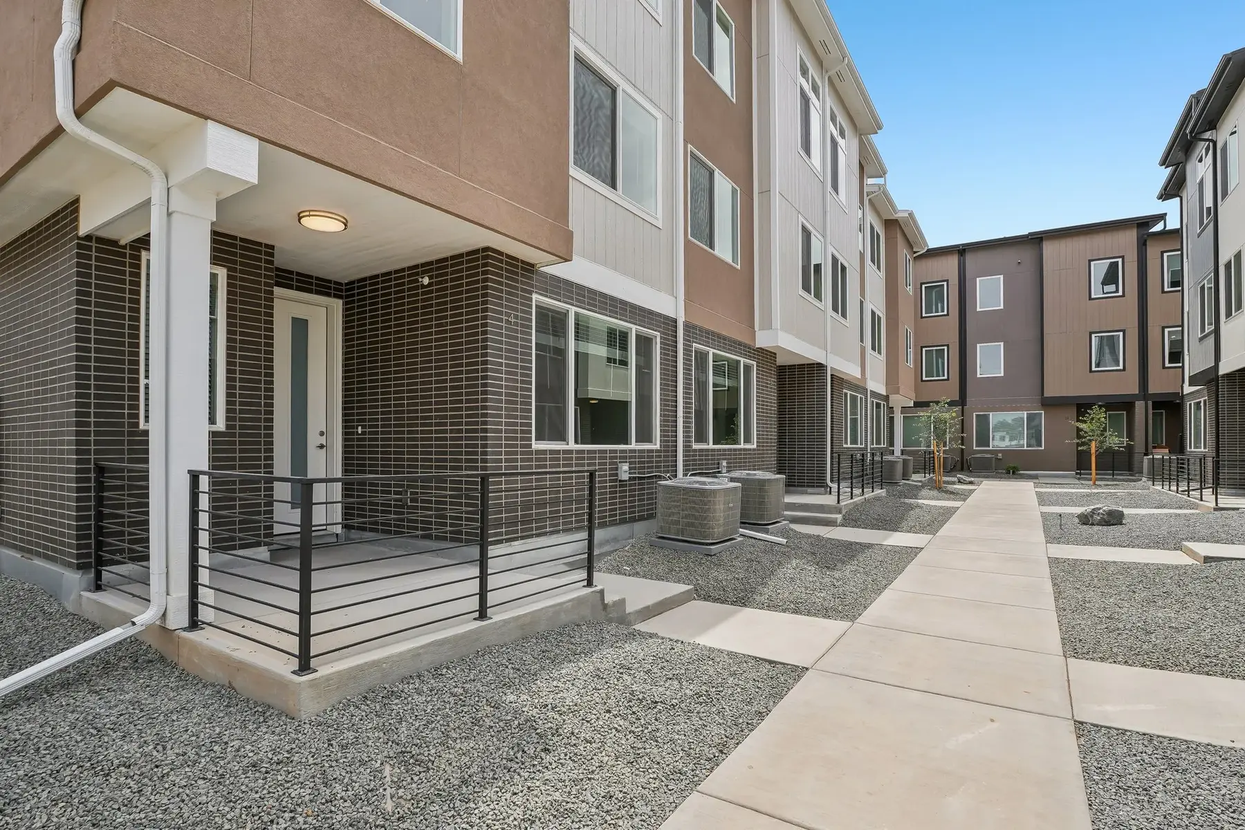 A modern outdoor area of an apartment complex featuring a pedestrian pathway and structured buildings with a mix of brown and beige siding. Graveled landscaping surrounds the entrance of the apartments and a clear blue sky is visible above.