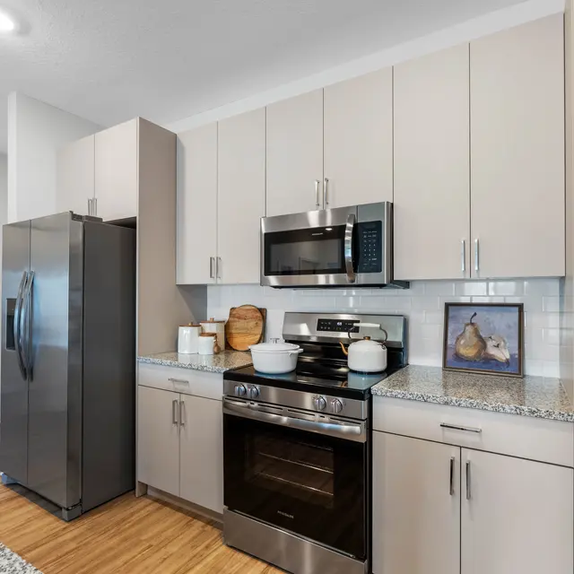 A sleek, modern kitchen featuring stainless steel appliances, including a refrigerator, oven, and microwave. The cabinets are light-colored with a granite countertop, and there is a framed artwork of pears on the counter.