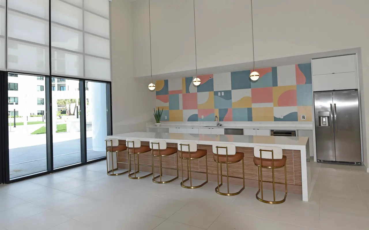 A bright, modern kitchen with a long white countertop and several brown bar stools. Behind the countertop is a colorful abstract wall design. Large glass doors lead outside, and a stainless steel refrigerator is visible on the right side.