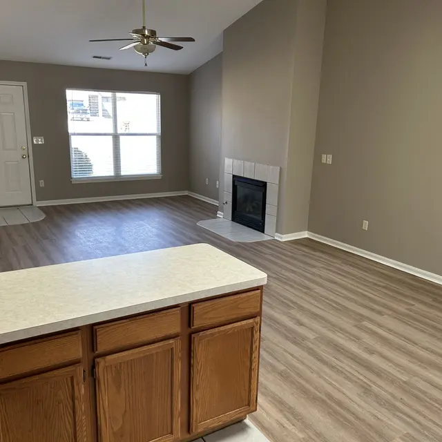 An empty living room with a fireplace and large window, featuring wooden cabinets and laminate flooring.