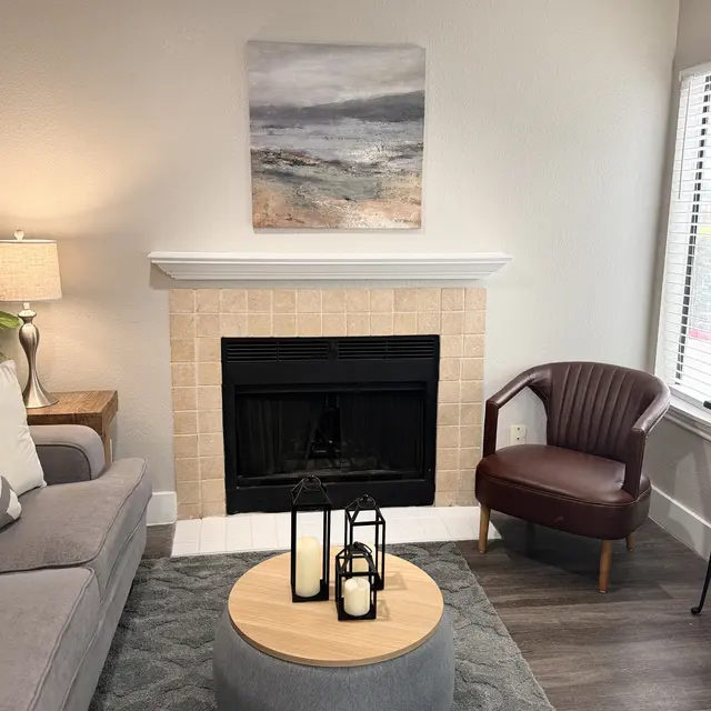 Cozy Living Room Design A cozy living room featuring a gray sofa, a round coffee table with candles, and a black fireplace. A painting of a seascape hangs above the fireplace. There is a brown armchair beside the fireplace and a lamp on a side table.
