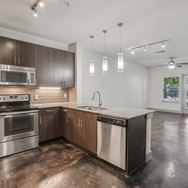 A modern kitchen with stainless steel appliances including an oven, microwave, and dishwasher. The kitchen features dark cabinetry and a gray countertop, with pendant lights hanging above. The adjacent living area has a ceiling fan and a door leading to an outdoor view.