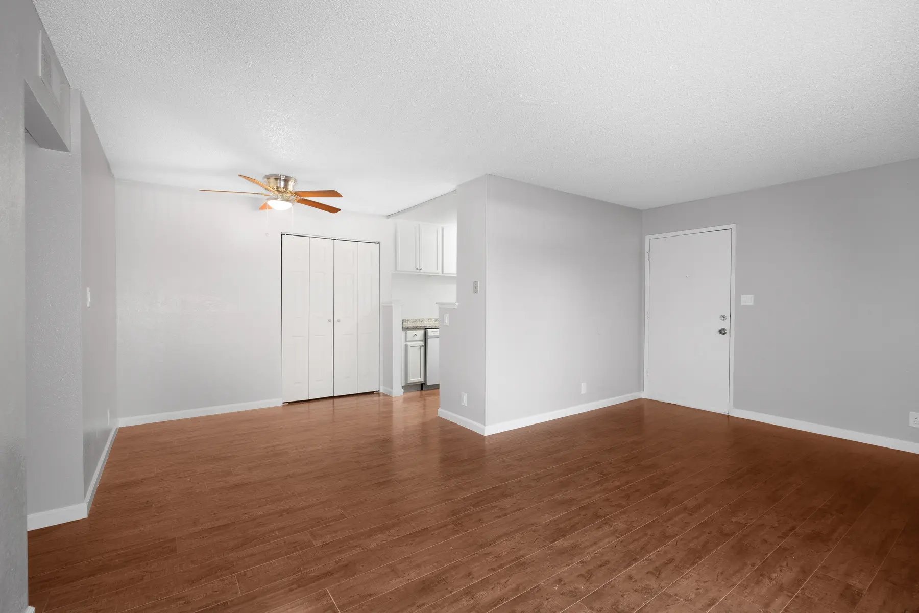 Spacious living room with light wood flooring and a ceiling fan, featuring a kitchen area to the left and an entrance door on the right.
