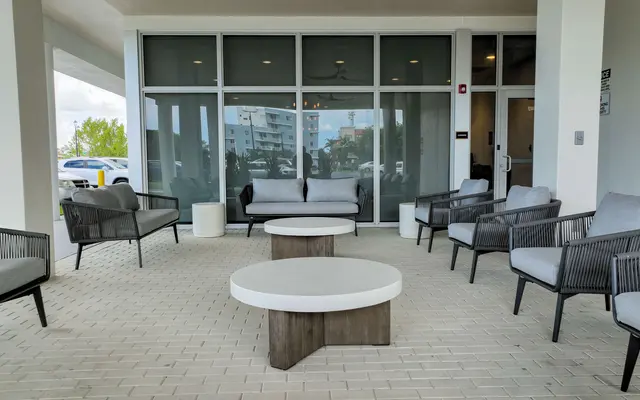 A modern outdoor lounge area featuring several grey chairs and a round coffee table on a tiled surface. Glass windows provide a view of outside.