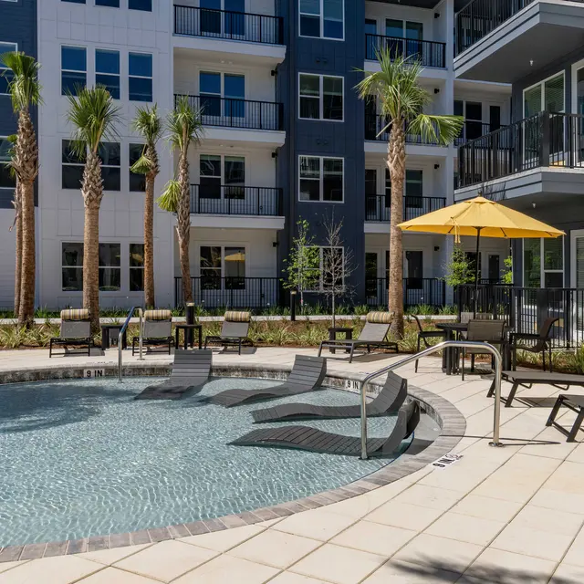 A modern apartment complex pool area featuring lounge chairs, palm trees, and an umbrella.