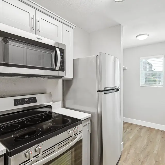 A modern kitchen featuring stainless steel appliances including a microwave, oven, and refrigerator. The space has white cabinetry and a light-colored countertop, with a window providing natural light.