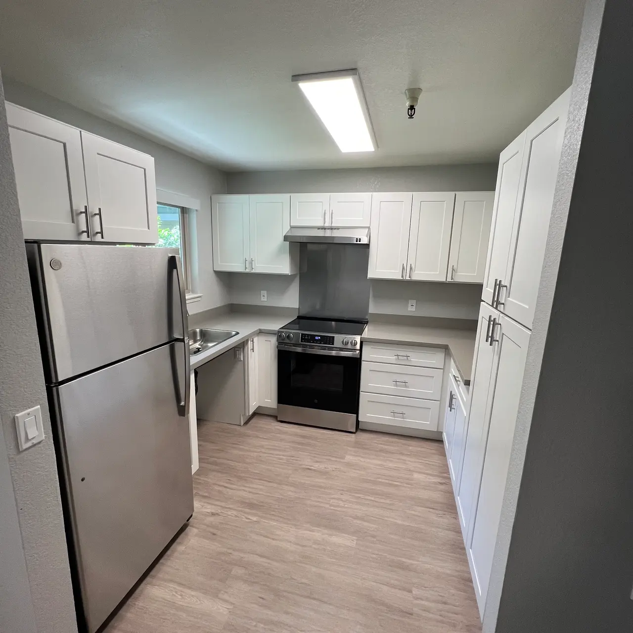 A modern kitchen featuring white cabinets, stainless steel appliances including a refrigerator and oven, and a light wood floor. The layout is spacious, with good lighting from a window and a ceiling fixture.