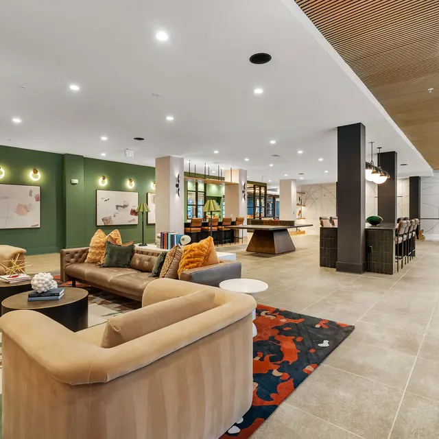 A spacious modern lounge area featuring plush seating in warm tones, decorative rugs, and contemporary lighting. The room is divided by decorative columns, with a bar-style table visible in the background alongside dining chairs. Green accent walls add vibrancy to the decor.