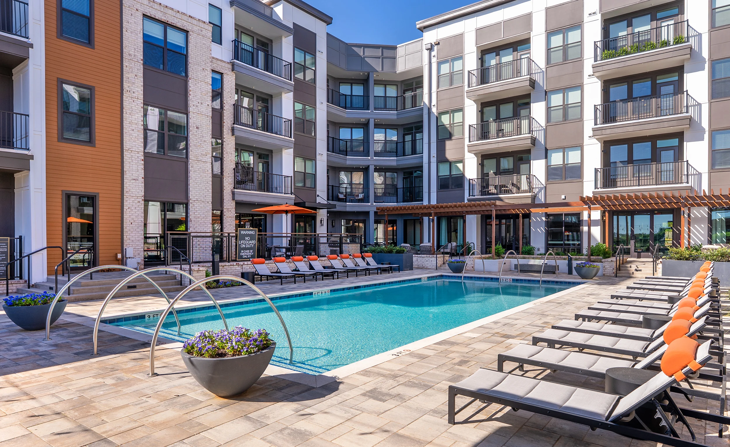 A modern apartment complex featuring a swimming pool surrounded by lounge chairs and planters.