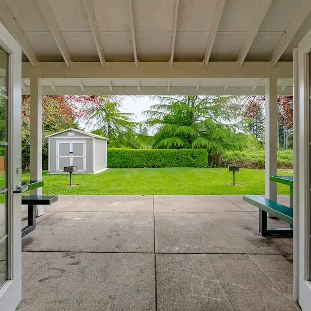 View from a covered patio looking out towards a grassy area with a small shed and grill stations, framed by white double doors.