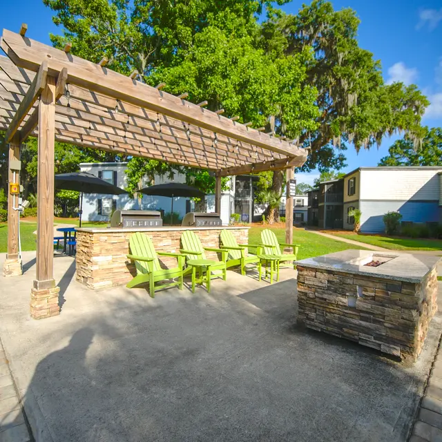 A beautifully landscaped outdoor gathering area featuring a wooden pergola and stone fire pit, surrounded by green chairs and residential buildings.
