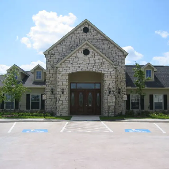A large building with a stone facade and a triangular roof, featuring a main entrance with double wooden doors and several windows. The building has a well-maintained landscape with grass and trees, and parking spaces marked in front.