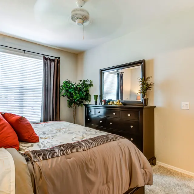 A cozy bedroom featuring a bed with red and beige bedding, a dark wooden dresser, a mirror, and large windows with curtains. A green plant is placed beside the dresser, and the room is softly illuminated, suggesting a welcoming atmosphere.