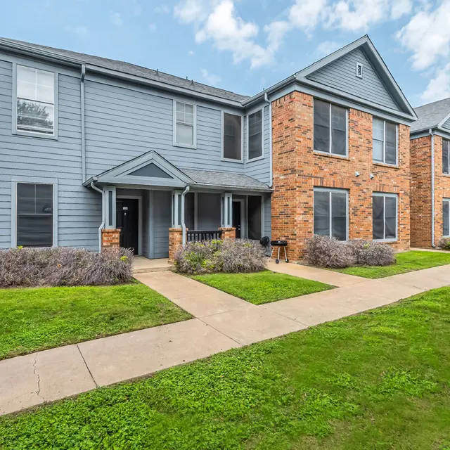 Exterior view of a two-story apartment complex with gray siding and brick accents, surrounded by a grassy area and walkway.